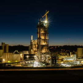 Das Bild zeigt eine Industrieanlage bei Nacht mit mehreren beleuchteten Gebäuden und Strukturen. Im Zentrum steht ein hoher Turm mit einem Kran an der Spitze, von dem helles Licht ausgeht. Umgeben ist der Turm von zylindrischen Tanks und weiteren technischen Anlagen, im Hintergrund ist ein dunkler Himmel mit leichtem Dämmerlicht am Horizont zu sehen.