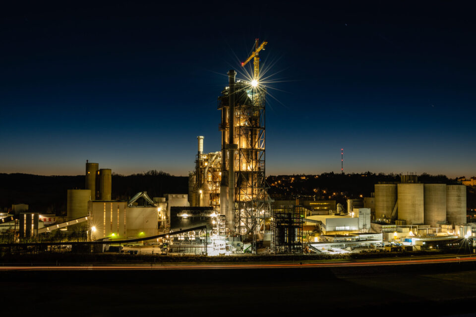 Das Bild zeigt eine Industrieanlage bei Nacht mit mehreren beleuchteten Gebäuden und Strukturen. Im Zentrum steht ein hoher Turm mit einem Kran an der Spitze, von dem helles Licht ausgeht. Umgeben ist der Turm von zylindrischen Tanks und weiteren technischen Anlagen, im Hintergrund ist ein dunkler Himmel mit leichtem Dämmerlicht am Horizont zu sehen.