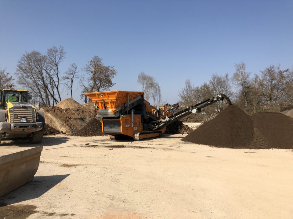 Auf dem Bild ist eine Baustelle mit einem großen orangefarbenen Gerät in der Mitte zu sehen, vermutlich ein Sieb- oder Brecher für Erdmaterial. Um das Gerät herum liegen mehrere Haufen aus Erde und Kies. Links im Bild steht ein gelbes Baustellenfahrzeug, wahrscheinlich ein Bulldozer oder Radlader, im Hintergrund sind kahle Bäume und ein klarer blauer Himmel zu erkennen.