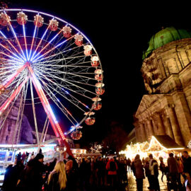 Das Bild zeigt eine lebhafte nächtliche Szene auf einem festlichen Jahrmarkt mit einem großen, bunt beleuchteten Riesenrad im Mittelpunkt. Zahlreiche Menschen sind auf dem Platz unterwegs, und rechts neben dem Riesenrad steht ein prunkvolles Gebäude mit grünlicher Kuppel. Mehrere kleine, geschmückte Stände deuten auf einen Weihnachtsmarkt oder ein ähnliches Fest hin.