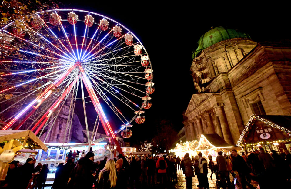 Das Bild zeigt eine lebhafte nächtliche Szene auf einem festlichen Jahrmarkt mit einem großen, bunt beleuchteten Riesenrad im Mittelpunkt. Zahlreiche Menschen sind auf dem Platz unterwegs, und rechts neben dem Riesenrad steht ein prunkvolles Gebäude mit grünlicher Kuppel. Mehrere kleine, geschmückte Stände deuten auf einen Weihnachtsmarkt oder ein ähnliches Fest hin.
