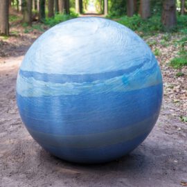 Das Bild zeigt eine große Kugel mit blau-weißer Musterung, die an einen Planeten oder eine abstrakte Darstellung von Wasser und Himmel erinnert. Sie liegt auf einem unbefestigten Weg in einem bewaldeten Gebiet mit grünem Laub und Sonnenlicht, das durch die Bäume fällt.