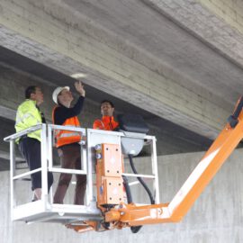 Das Bild zeigt drei Personen in einer hydraulischen Hebebühne, die die Unterseite einer Betonbrücke oder eines Überführungsbauwerks inspizieren. Alle tragen Sicherheitswesten und Helme, eine Person zeigt auf die Decke der Konstruktion. Die Hebebühne ist orange und mit sichtbaren Bedienelementen ausgestattet.