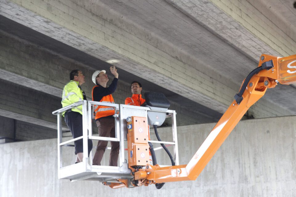 Das Bild zeigt drei Personen in einer hydraulischen Hebebühne, die die Unterseite einer Betonbrücke oder eines Überführungsbauwerks inspizieren. Alle tragen Sicherheitswesten und Helme, eine Person zeigt auf die Decke der Konstruktion. Die Hebebühne ist orange und mit sichtbaren Bedienelementen ausgestattet.