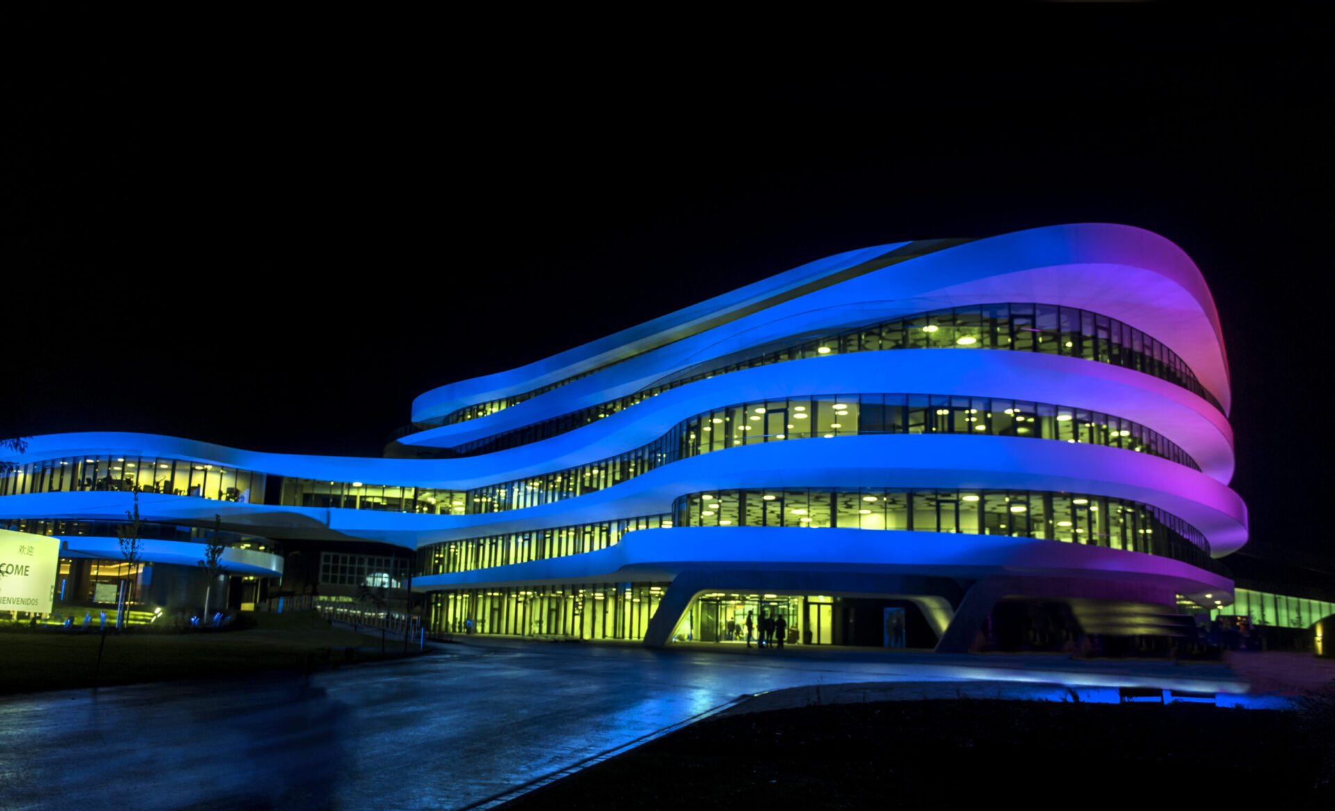 Das Bild zeigt ein modernes, mehrstöckiges Gebäude mit einer wellenförmigen Architektur. In der Nachtaufnahme wird das Gebäude durch blaues und violettes Licht beleuchtet, wodurch die geschwungene Struktur und die großen Fenster besonders hervorgehoben werden.