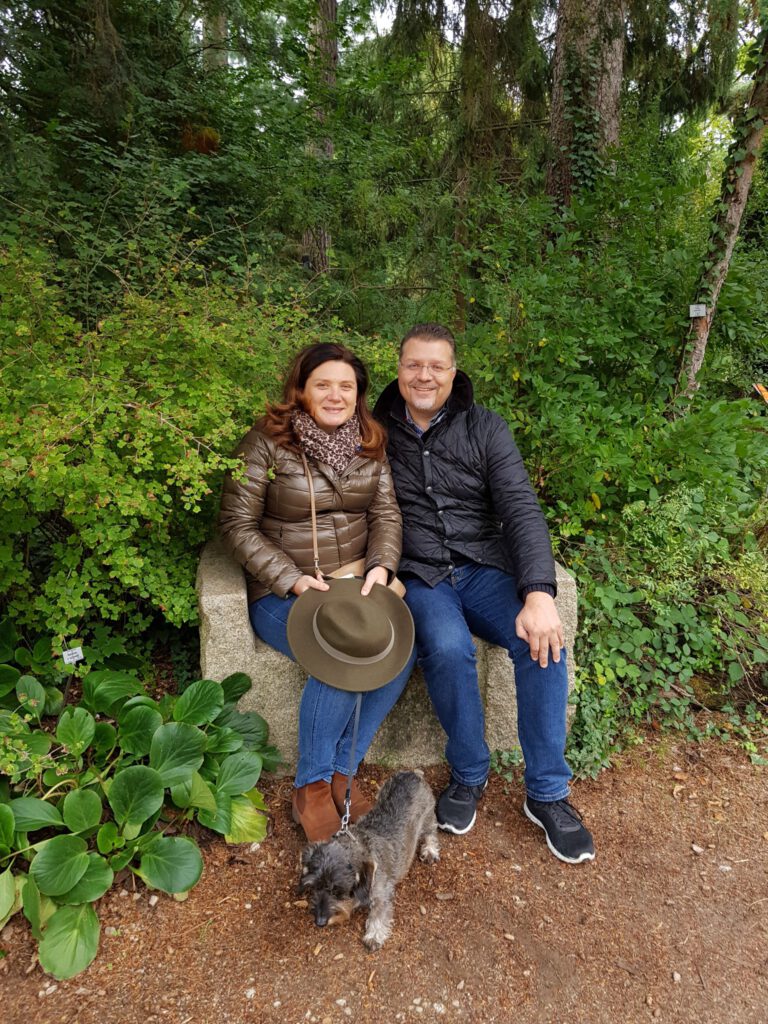 Das Bild zeigt zwei Personen, die auf einer Steinbank in einem grünen, bewaldeten Gebiet sitzen. Beide tragen Jacken und Jeans, eine Person hält einen Hut im Schoß. Vor ihnen steht ein kleiner angeleinter Hund auf dem Boden.