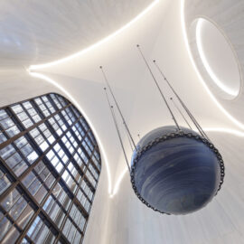 Das Bild zeigt einen großen, modernen Innenraum mit hoher Decke und geschwungenem Design mit eingelassener Beleuchtung. In der Mitte hängt eine kugelförmige Skulptur, die an einen Himmelskörper erinnert, an mehreren Ketten von der Decke. Eine große Fensterfläche mit Gitterstruktur lässt Tageslicht in den Raum.