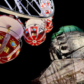 Rote Gondeln eines Riesenrads. Rechts neben dem Riesenrad steht ein prunkvolles Gebäude mit grünlicher Kuppel.