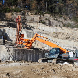 Das Bild zeigt eine Baustelle mit einem großen orangefarbenen Bohrgerät auf felsigem Untergrund. Eine Person steht in der Nähe der Maschine, die in eine Felswand bohrt. Im Hintergrund sind terrassenartige Felsformationen und Bäume am oberen Rand des Hangs zu sehen.