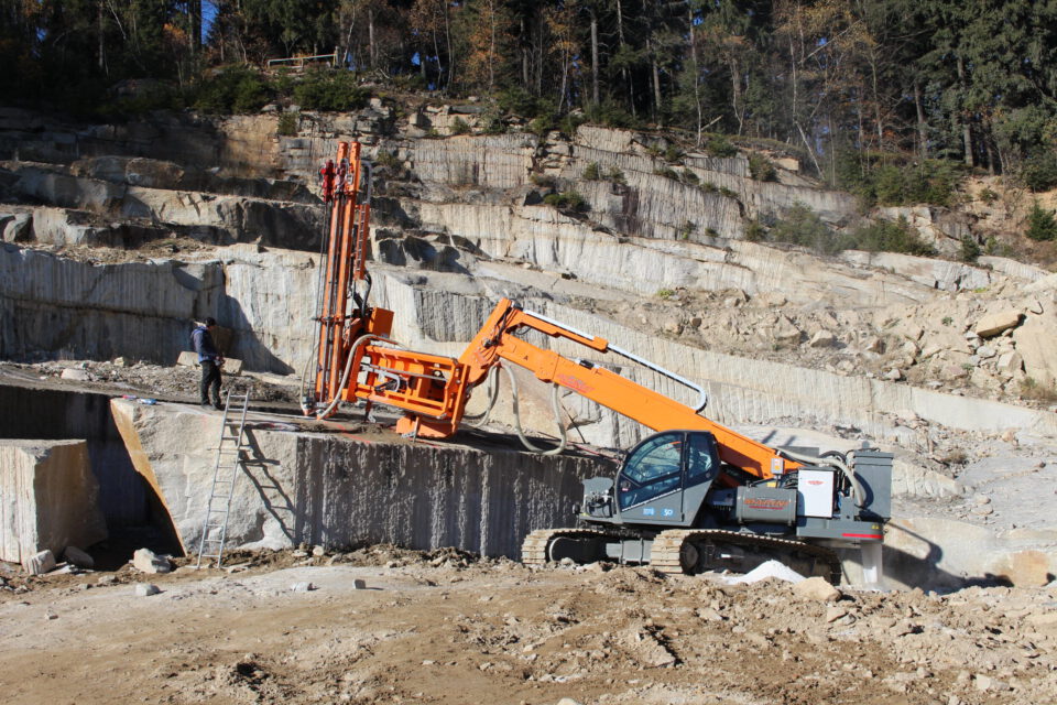 Das Bild zeigt eine Baustelle mit einem großen orangefarbenen Bohrgerät auf felsigem Untergrund. Eine Person steht in der Nähe der Maschine, die in eine Felswand bohrt. Im Hintergrund sind terrassenartige Felsformationen und Bäume am oberen Rand des Hangs zu sehen.