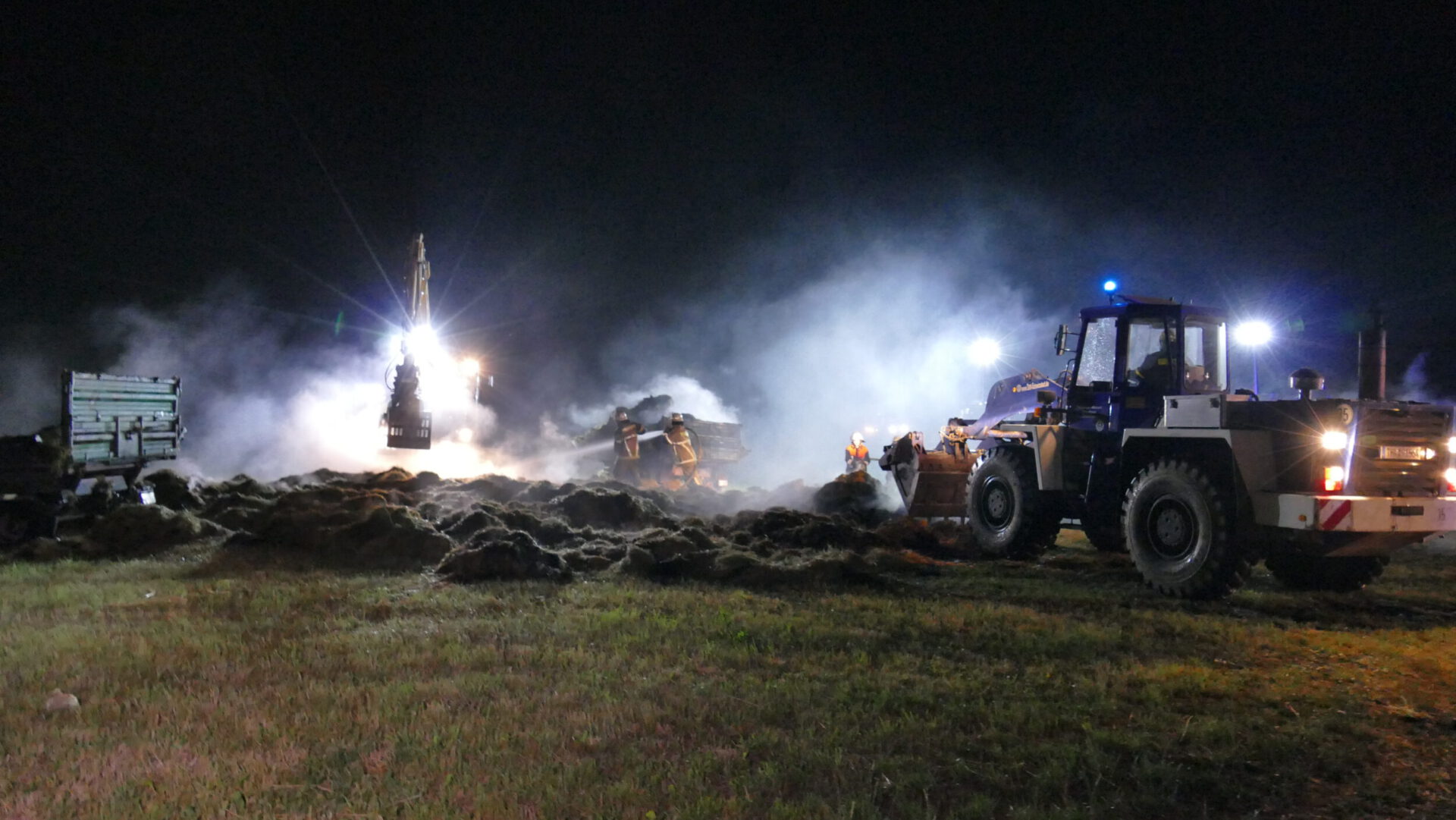 Das Bild zeigt eine nächtliche Einsatzszene auf einem Feld mit schwerem Gerät und mehreren Arbeitern. Im Vordergrund steht ein großes Fahrzeug mit eingeschaltetem Scheinwerfer, während im Hintergrund weitere Maschinen und Fahrzeuge zu sehen sind. Rauch oder Nebel wird von hellem Licht durchleuchtet, und Personen in reflektierender Schutzkleidung sind im Einsatz.