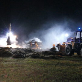 Das Bild zeigt eine nächtliche Einsatzszene auf einem Feld mit schwerem Gerät und mehreren Arbeitern. Im Vordergrund steht ein großes Fahrzeug mit eingeschaltetem Scheinwerfer, während im Hintergrund weitere Maschinen und Fahrzeuge zu sehen sind. Rauch oder Nebel wird von hellem Licht durchleuchtet, und Personen in reflektierender Schutzkleidung sind im Einsatz.