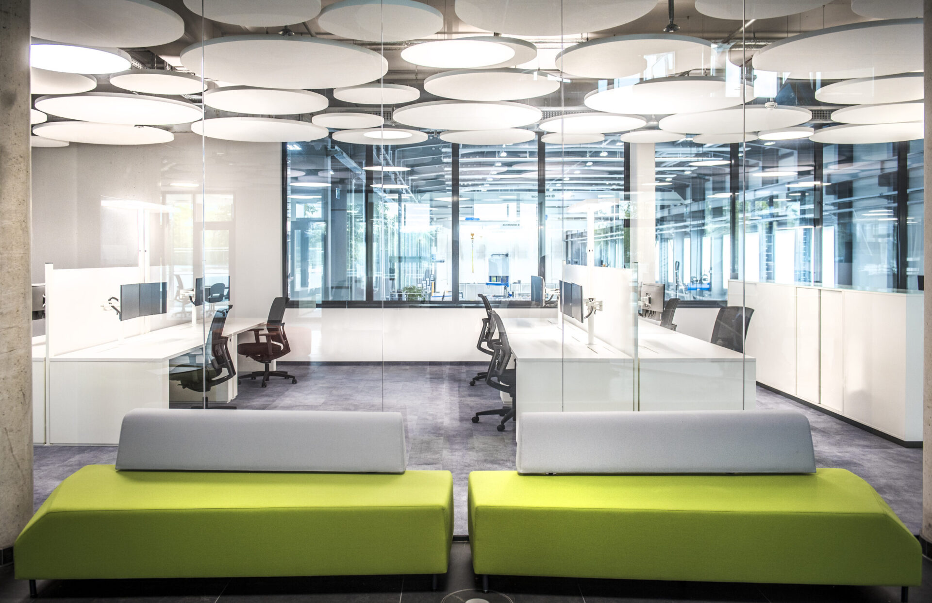 Das Bild zeigt ein modernes Büro mit einer Glaswand, die zwei Bereiche voneinander trennt. Im Vordergrund stehen ein hellgraues und ein leuchtend grünes Sofa. Hinter der Glaswand befinden sich mehrere Arbeitsplätze mit Stühlen und Monitoren, und die Decke ist mit runden Akustikpaneelen ausgestattet.