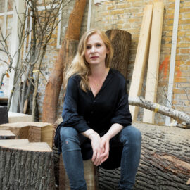 Das Bild zeigt eine Person, die auf einem Holzstamm in einer Werkstatt oder einem Atelier sitzt, umgeben von weiteren Holzstücken und Baumstämmen. Im Hintergrund sind eine Ziegelwand und einige Äste zu sehen.