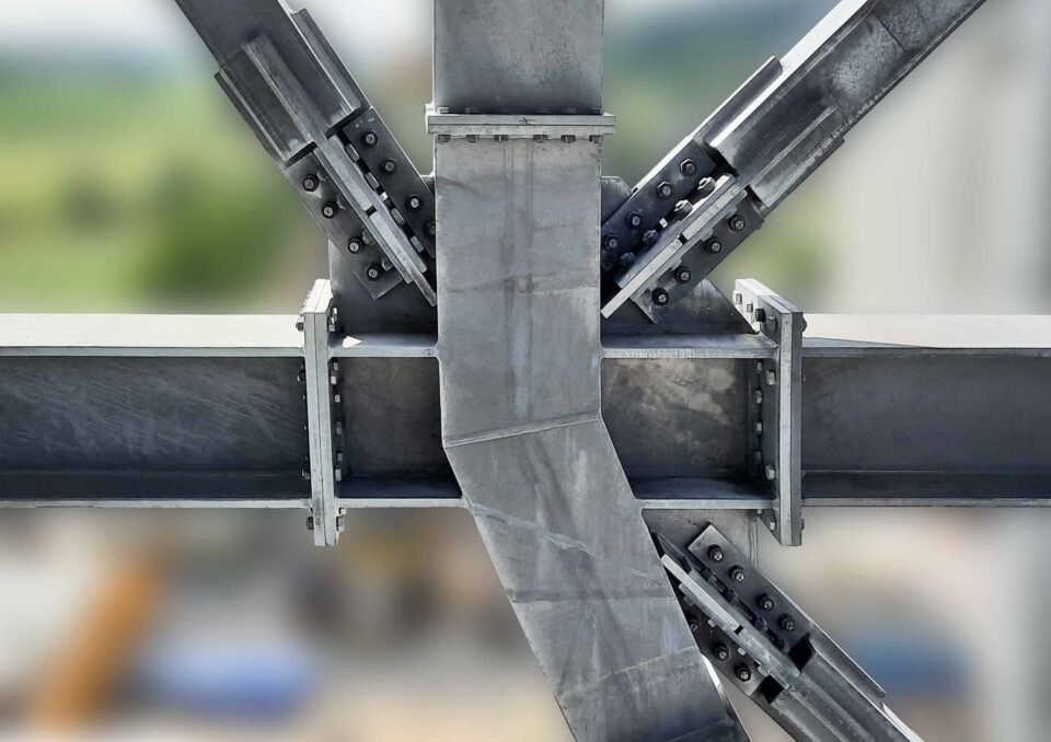 Das Bild zeigt eine Nahaufnahme eines Stahlverbindungspunkts, an dem mehrere Stahlträger mit zahlreichen Schrauben und Platten miteinander verbunden sind. Die Konstruktion bildet eine stabile und tragfähige Verbindung. Der Hintergrund ist unscharf, wodurch der Fokus auf dem Verbindungspunkt liegt.