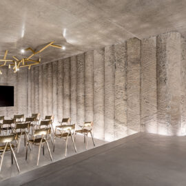 Das Bild zeigt einen modernen, minimalistisch gestalteten Raum mit Betonwänden und -boden. Mehrere Reihen goldfarbener Stühle sind in Richtung eines großen Flachbildschirms aufgestellt, der auf einem Ständer montiert ist. An der Decke befindet sich eine kunstvolle Leuchte aus unregelmäßig angeordneten goldenen Stäben, und die Wände weisen ein strukturiertes Muster auf.
