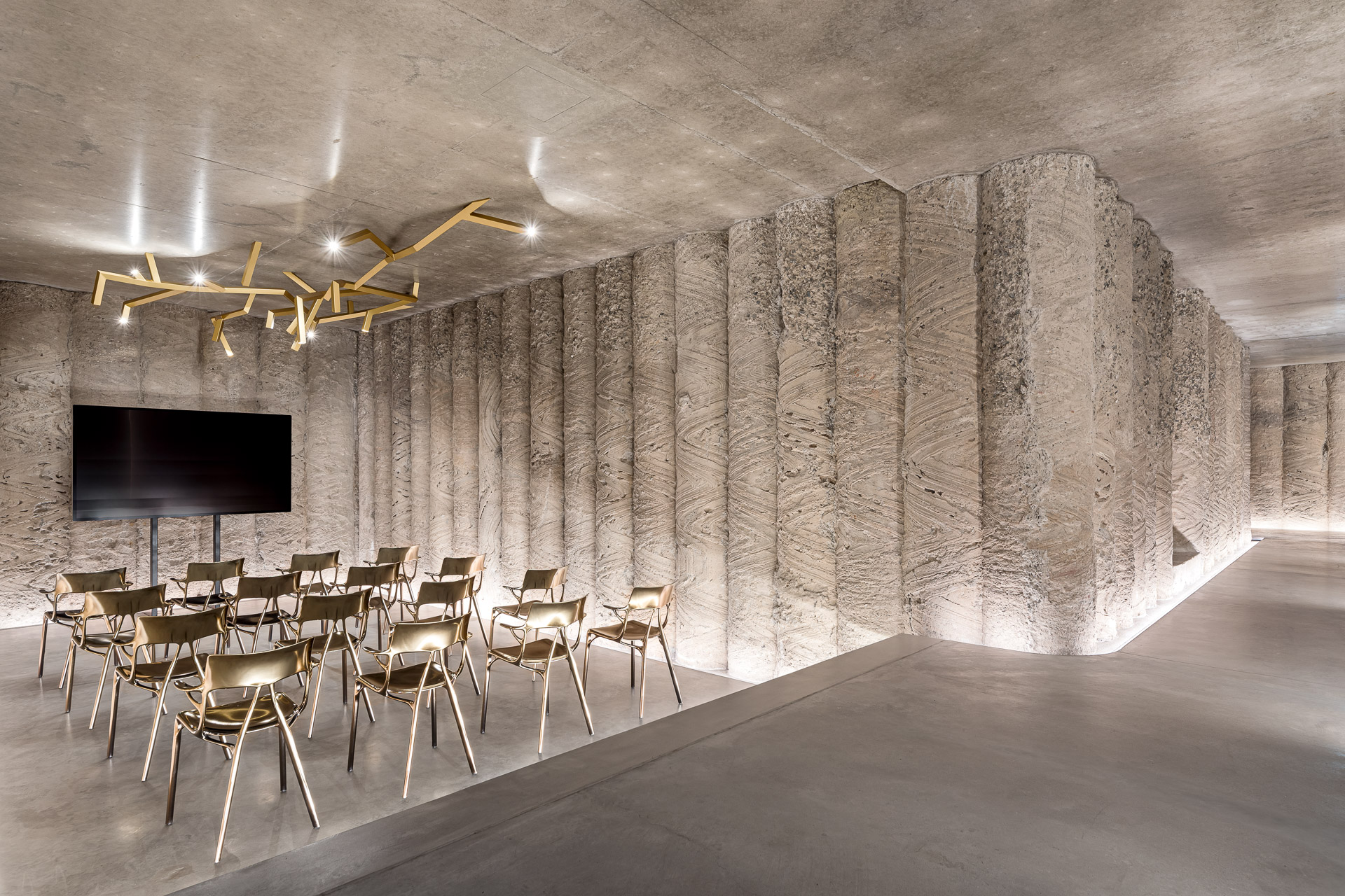 Das Bild zeigt einen modernen, minimalistisch gestalteten Raum mit Betonwänden und -boden. Mehrere Reihen goldfarbener Stühle sind in Richtung eines großen Flachbildschirms aufgestellt, der auf einem Ständer montiert ist. An der Decke befindet sich eine kunstvolle Leuchte aus unregelmäßig angeordneten goldenen Stäben, und die Wände weisen ein strukturiertes Muster auf.