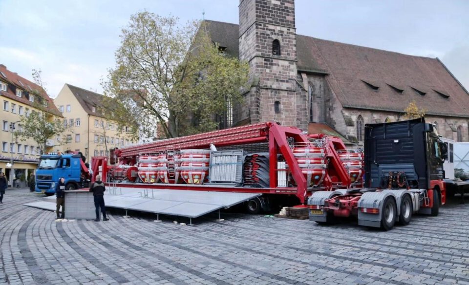 Das zweite Bild zeigt den Aufbau eines großen rot-weißen Fahrgeschäfts auf einem gepflasterten Platz in einer Stadt. Das Fahrgeschäft befindet sich noch auf einem Anhänger, der an einen LKW gekoppelt ist. Mehrere Personen arbeiten rund um das Fahrgeschäft, vermutlich beim Aufbau. Im Hintergrund sind Gebäude und eine Kirche zu sehen, was auf eine zentrale Lage – möglicherweise ein Marktplatz – hinweist.