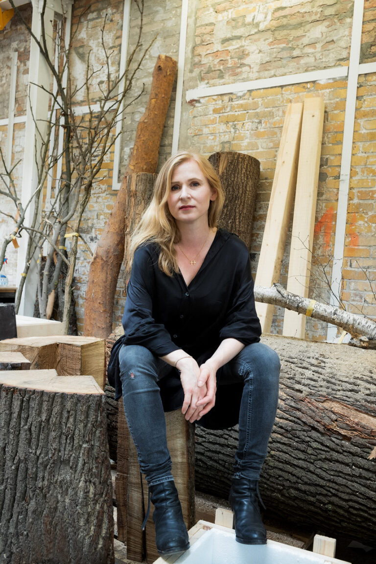 Das Bild zeigt eine Person, die auf einem Holzstamm in einer Werkstatt oder einem Atelier sitzt, umgeben von weiteren Holzstücken und Baumstämmen. Im Hintergrund sind eine Ziegelwand und einige Äste zu sehen.