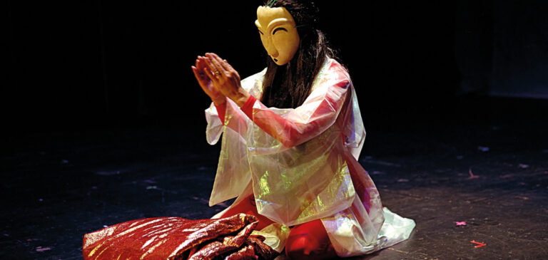 Das Bild zeigt eine Person mit einer Maske, die auf einer Bühne kniet und ein glänzendes, durchscheinendes Gewand mit langen Ärmeln trägt. Die Hände sind vor dem Gesicht zusammengelegt, und über den Knien liegt ein roter und goldener Stoff. Der Hintergrund ist dunkel, was auf eine Aufführung oder Präsentation in gedämpfter Beleuchtung hindeutet.