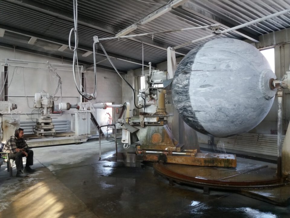 Das Bild zeigt eine industrielle Umgebung mit einer großen Kugel aus Stein oder ähnlichem Material, die auf einer rotierenden Plattform bearbeitet wird. Um die Kugel herum befinden sich mehrere mechanische Arme und Geräte zur Bearbeitung. Im Hintergrund sitzt eine Person auf einem Stuhl und beobachtet den Vorgang in einer Werkstatt mit Betonboden und Metallbedachung.