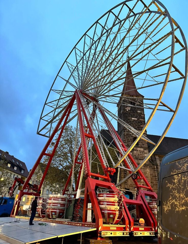 Ein großes Riesenrad wird in einer städtischen Umgebung aufgebaut oder abgebaut. Die Metallstruktur ist teilweise errichtet und wird von roten Trägern gestützt, während mehrere Personen am Boden arbeiten. Im Hintergrund sind eine Kirche mit hohem Turm und weitere Gebäude zu sehen, der Himmel ist in einem blauen Ton gehalten.