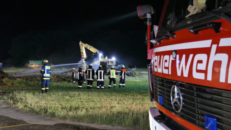 Das Bild zeigt eine nächtliche Einsatzszene der Feuerwehr auf einem grasbewachsenen Feld. Im Vordergrund steht ein rotes Feuerwehrfahrzeug, mehrere Feuerwehrleute in Schutzkleidung sind im Einsatz. Zwei Feuerwehrleute sprühen Wasser, während helle Scheinwerfer die Umgebung ausleuchten.