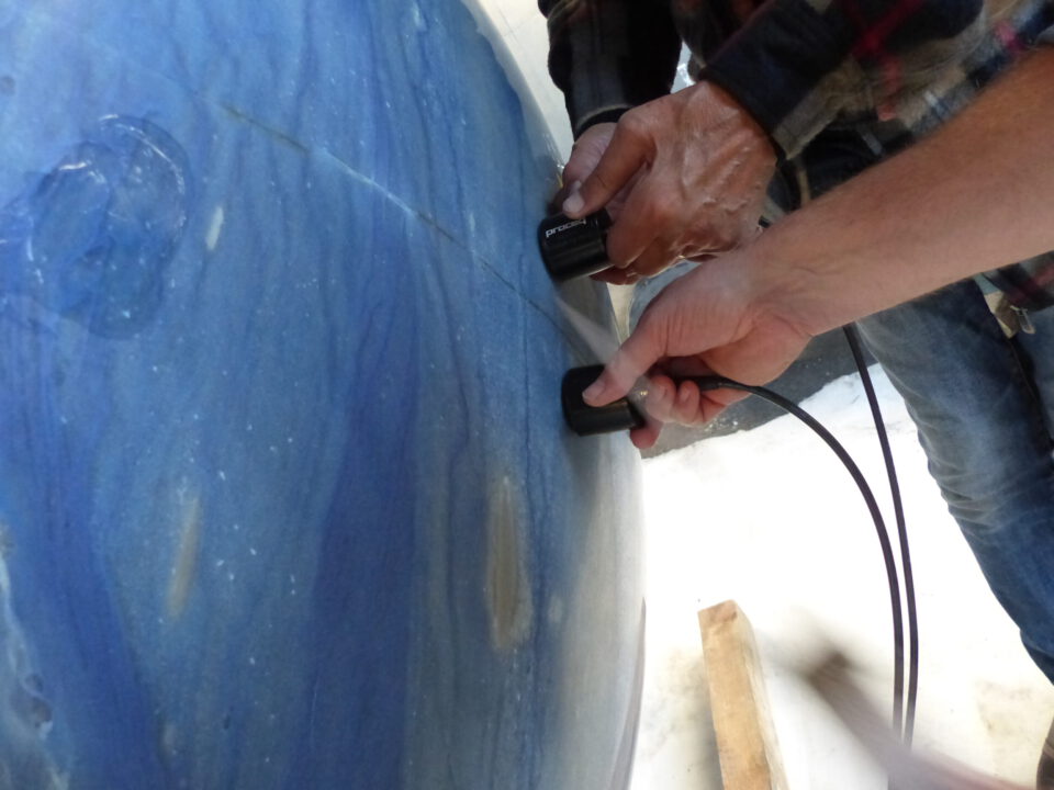 Das Bild zeigt zwei Personen, die mit Handgeräten die Oberfläche einer großen, blauen Kugel untersuchen oder vermessen. Die Kugel hat eine glatte Oberfläche mit helleren und dunkleren blauen Streifen. Nur die Hände der Personen sind sichtbar.