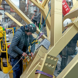 Das Bild zeigt zwei Arbeiter mit Sicherheitsausrüstung, darunter Helme und Gurte, bei der Arbeit an einer großen Metallkonstruktion mit gelben Trägern. Einer der Arbeiter bedient ein Werkzeug, um eine Kette an der Struktur zu befestigen oder zu justieren. Im Hintergrund sind Gebäude mit traditioneller Architektur sowie ein Baugerüst zu sehen.