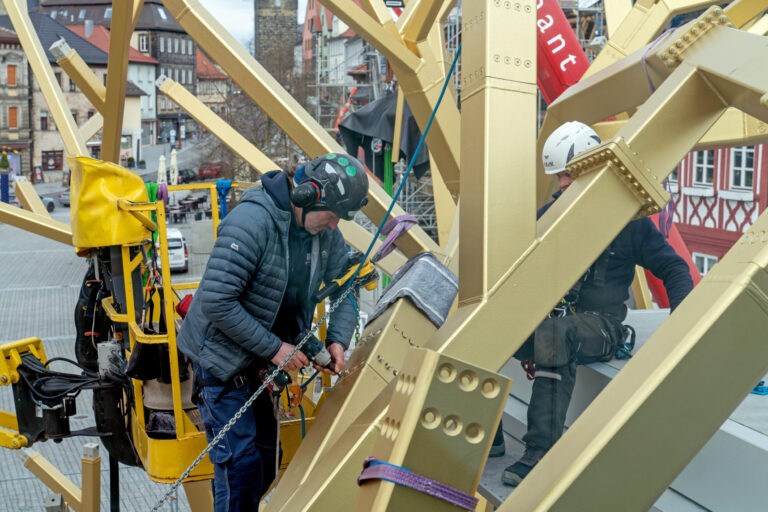 Das Bild zeigt zwei Arbeiter mit Sicherheitsausrüstung, darunter Helme und Gurte, bei der Arbeit an einer großen Metallkonstruktion mit gelben Trägern. Einer der Arbeiter bedient ein Werkzeug, um eine Kette an der Struktur zu befestigen oder zu justieren. Im Hintergrund sind Gebäude mit traditioneller Architektur sowie ein Baugerüst zu sehen.