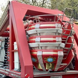 Das Bild zeigt eine Nahaufnahme von rot-weißen Sitzkabinen eines Fahrgeschäfts, vermutlich eines Riesenrads, die auf einem Transportanhänger gestapelt sind. Im Hintergrund sind Gebäude und Bäume zu erkennen.