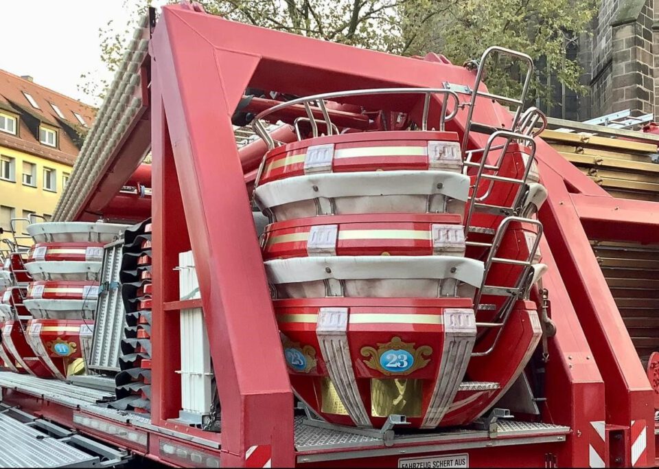 Das Bild zeigt eine Nahaufnahme von rot-weißen Sitzkabinen eines Fahrgeschäfts, vermutlich eines Riesenrads, die auf einem Transportanhänger gestapelt sind. Im Hintergrund sind Gebäude und Bäume zu erkennen.