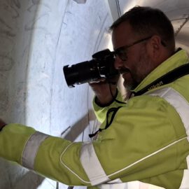 Eine Person mit einer Warnjacke fotografiert im Inneren eines Tunnels eine Stelle an der Tunnelwand. Die Person hält eine Kamera in der Hand und dokumentiert oder inspiziert vermutlich die Oberfläche. Das Gesicht der Person ist unkenntlich gemacht.