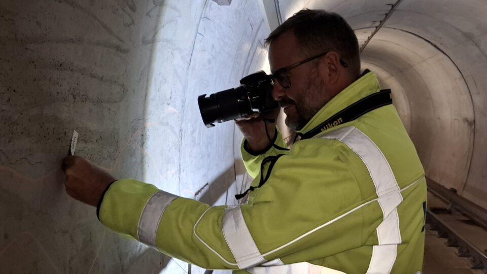 Eine Person mit einer Warnjacke fotografiert im Inneren eines Tunnels eine Stelle an der Tunnelwand. Die Person hält eine Kamera in der Hand und dokumentiert oder inspiziert vermutlich die Oberfläche. Das Gesicht der Person ist unkenntlich gemacht.