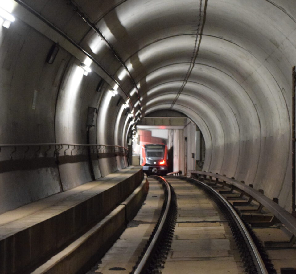 Das Bild zeigt einen U-Bahn-Tunnel, in dem ein Zug aus der Ferne heranfährt. Der Tunnel ist gut beleuchtet, mit einer begehbaren Fläche links neben den Gleisen und gebogenen Betonwänden.