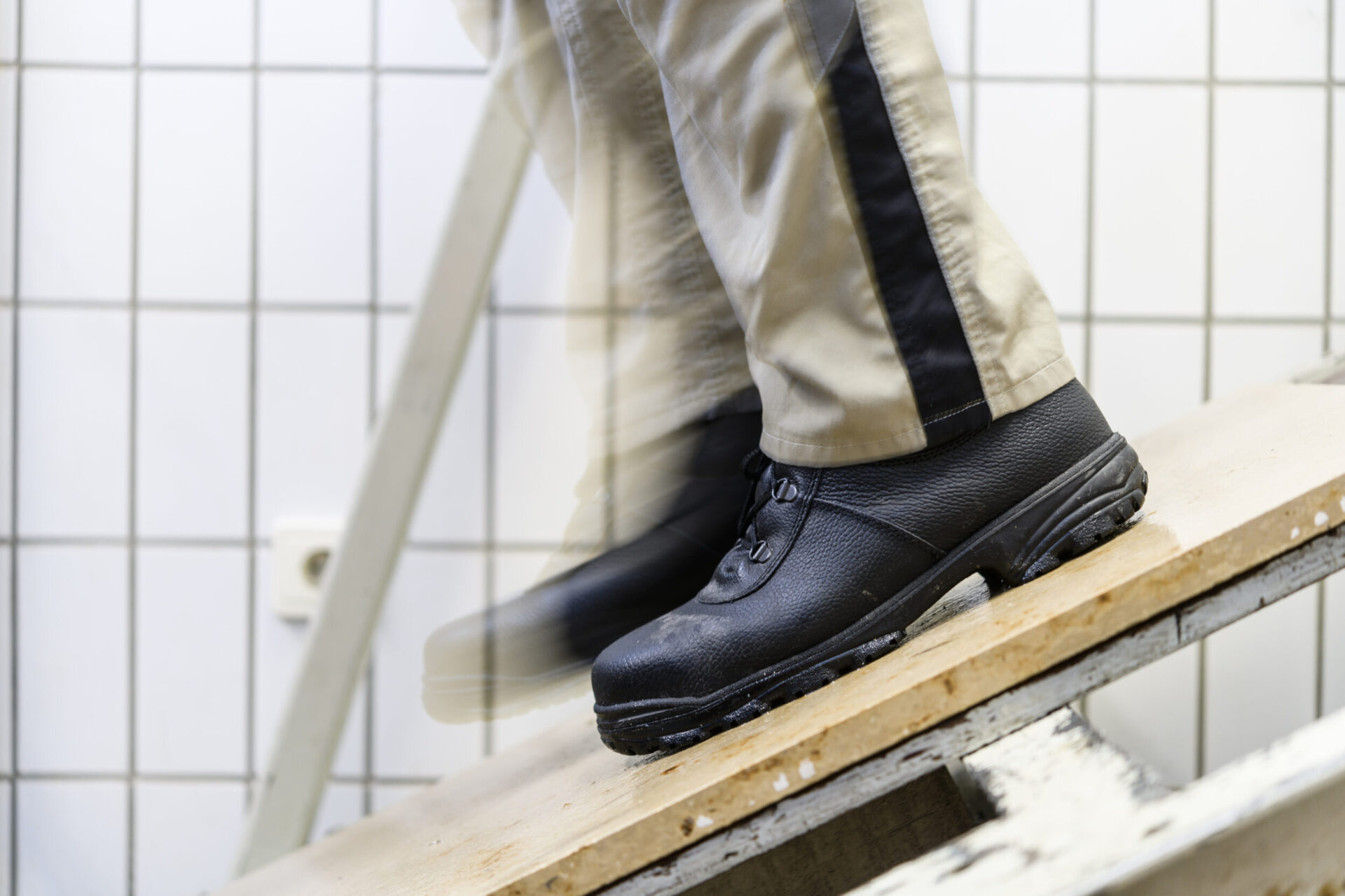 Das Bild zeigt eine Person mit schwarzen Arbeitsschuhen und beigefarbener Hose mit schwarzem Seitenstreifen, die über ein Holzbrett läuft. Der Hintergrund besteht aus weißen Fliesen mit einem Gittermuster. Eine Bewegungsunschärfe deutet auf eine laufende Bewegung hin.