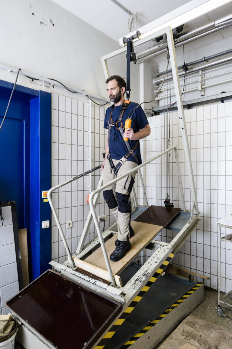 Das Bild zeigt eine Person, die auf einem schräg gestellten Hebebühne steht, das Teil eines Labors oder Testaufbaus zu sein scheint. Die Person ist mit Sicherheitsgurten an einem Rahmen über dem Gerät gesichert. Im Hintergrund sind geflieste Wände, Kabel und technische Geräte zu sehen, die auf eine kontrollierte Versuchsumgebung hinweisen.