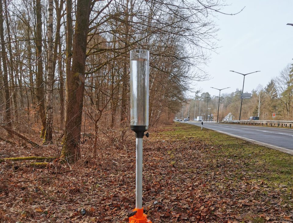 Das Bild zeigt einen Regenmesser, der auf einem Stativ mit drei Beinen und orangefarbenen Verbindungsstücken montiert ist. Der transparente Zylinder des Messgeräts steht auf einem Waldboden, der mit trockenen Blättern bedeckt ist. Im Hintergrund sind eine Straße mit Straßenlaternen und laublose Bäume zu sehen.