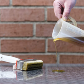 Das Bild zeigt eine Hand, die eine Kunststoff-Messkanne hält und eine gelbliche Flüssigkeit auf eine reflektierende Oberfläche gießt. Daneben liegt ein Pinsel mit weißem Griff, dessen Borsten ebenfalls mit der Flüssigkeit benetzt sind. Im Hintergrund sind rote Ziegelwände mit sichtbaren Mörtelfugen zu sehen.