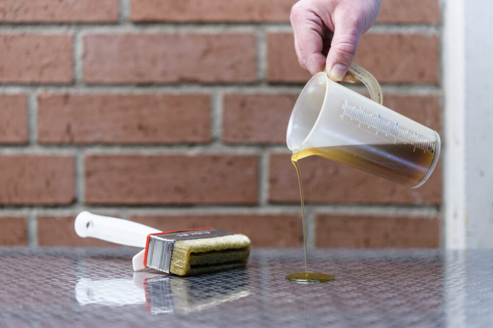 Das Bild zeigt eine Hand, die eine Kunststoff-Messkanne hält und eine gelbliche Flüssigkeit auf eine reflektierende Oberfläche gießt. Daneben liegt ein Pinsel mit weißem Griff, dessen Borsten ebenfalls mit der Flüssigkeit benetzt sind. Im Hintergrund sind rote Ziegelwände mit sichtbaren Mörtelfugen zu sehen.