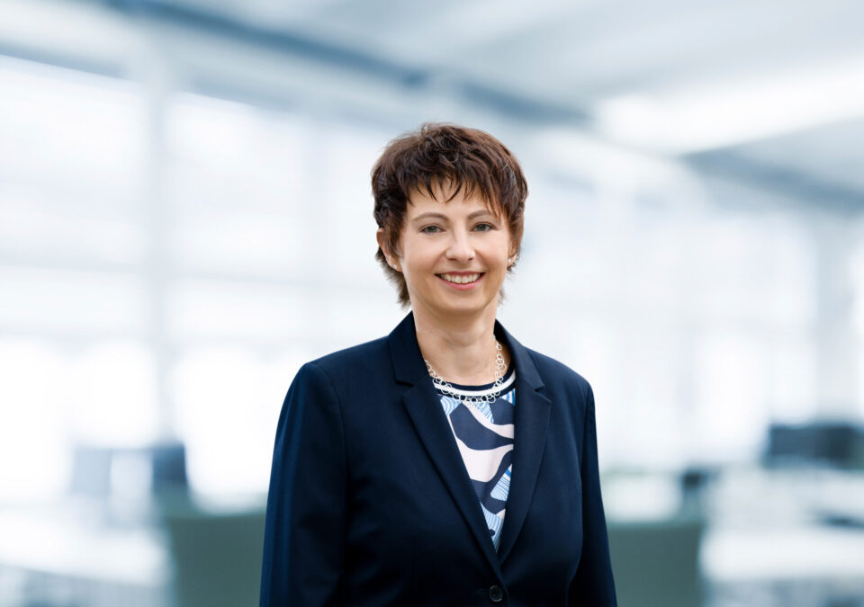 Das Bild zeigt eine Person in einem dunklen Blazer über einem gemusterten Hemd, stehend in einer Innenraumszene mit unscharfem Hintergrund, der auf eine Büroumgebung hinweist.