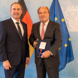 Zwei Männer im Anzug stehen nebeneinander. Der Mann rechts hält eine geöffnete Schachtel mit einer Medaille oder Auszeichnung. Im Hintergrund sind die Flaggen von Bayern, Deutschland und der Europäischen Union zu sehen.