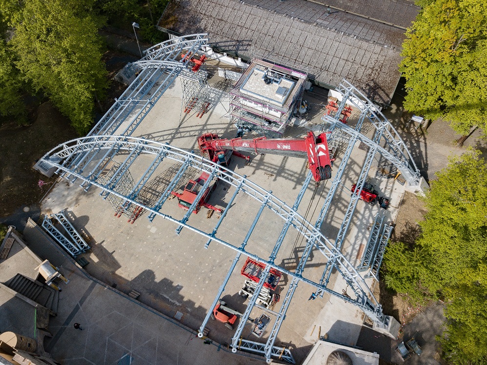 Das Bild zeigt eine Luftaufnahme einer Baustelle mit einer großen Metallkonstruktion, die das Gerüst eines Gebäudes oder Pavillons bildet. Innerhalb der Struktur befinden sich mehrere rote Baustellenfahrzeuge wie Kräne und Hebebühnen. Im Hintergrund sind Bäume und angrenzende Gebäude zu sehen.