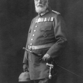 Das Bild zeigt eine historische Person in Militäruniform, die in der linken Hand einen Helm und in der rechten Hand ein Schwert hält. Auf der Brust sind mehrere Orden und Auszeichnungen zu sehen, darunter ein Eisernes Kreuz.