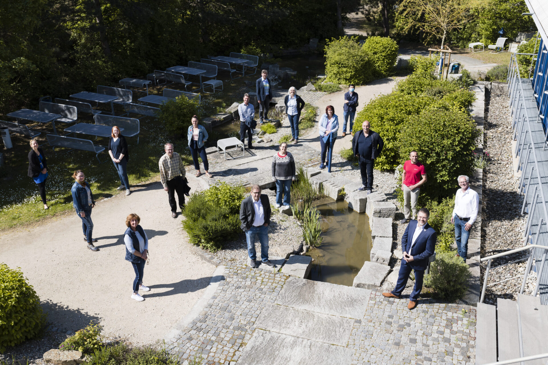 Das Bild zeigt eine Gruppe von Menschen, die im Freien in einer gartenähnlichen Umgebung stehen und Abstand zueinander halten. In der Mitte befindet sich ein kleines Wasserbecken mit Trittsteinen, umgeben von Pflanzen und Sträuchern. Im Hintergrund sind Tische, Bänke und ein Gebäude auf der rechten Seite zu sehen.