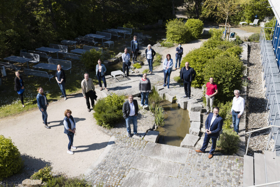 Das Bild zeigt eine Gruppe von Menschen, die im Freien in einer gartenähnlichen Umgebung stehen und Abstand zueinander halten. In der Mitte befindet sich ein kleines Wasserbecken mit Trittsteinen, umgeben von Pflanzen und Sträuchern. Im Hintergrund sind Tische, Bänke und ein Gebäude auf der rechten Seite zu sehen.