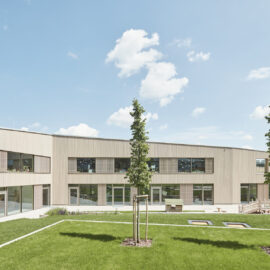 Das Bild zeigt ein modernes, eingeschossiges Gebäude mit heller Holzfassade und großen Fenstern und Türen. Davor befindet sich eine gepflegte Rasenfläche mit drei jungen Bäumen in einer Reihe. Im Hintergrund sind Hügel oder Berge unter einem leicht bewölkten Himmel zu sehen.