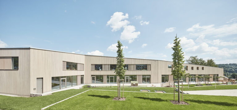 Das Bild zeigt ein modernes, eingeschossiges Gebäude mit heller Holzfassade und großen Fenstern und Türen. Davor befindet sich eine gepflegte Rasenfläche mit drei jungen Bäumen in einer Reihe. Im Hintergrund sind Hügel oder Berge unter einem leicht bewölkten Himmel zu sehen.