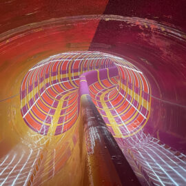 Das Bild zeigt das Innere einer bunten Wasserrutsche mit leuchtenden Streifen in Rot, Orange, Gelb und Lila. Die Oberfläche ist nass und glänzend, was auf eine Nutzung oder Betriebsbereitschaft hinweist.