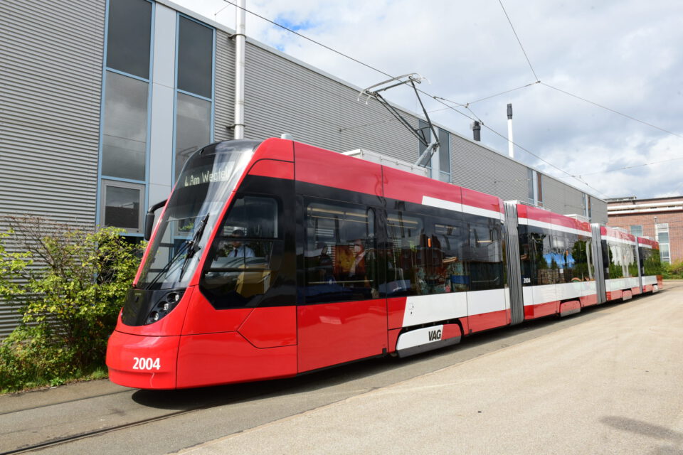 Ein moderner rot-weißer Straßenbahnwagen mit der Nummer 2004 steht auf einem Gleis neben einem großen Industriegebäude mit grauer Verkleidung und mehreren Fenstern. Die Straßenbahn ist über Oberleitungen elektrisch angeschlossen und zeigt die Liniennummer 4 mit dem Ziel „Am Wegfeld“. Die Umgebung wirkt wie ein Industrie- oder Wartungsbereich.